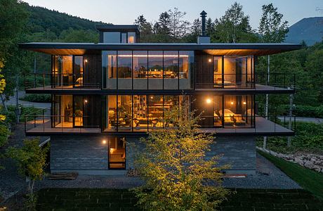 A modern, glass-walled house nestled in a forested landscape, with warm interior lighting.