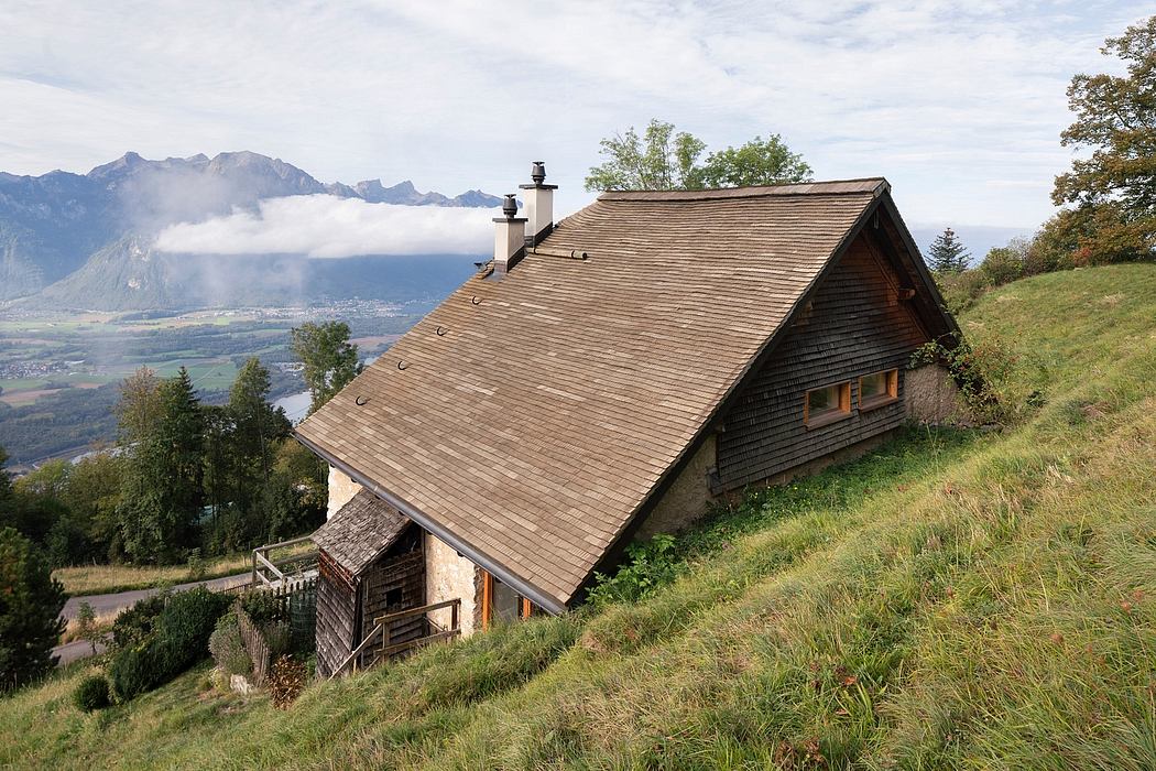 A rustic wooden chalet with a gabled roof and a balcony, set in a mountainous landscape.
