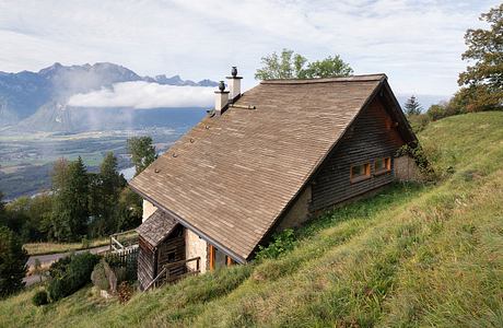 A rustic wooden chalet with a gabled roof and a balcony, set in a mountainous landscape.