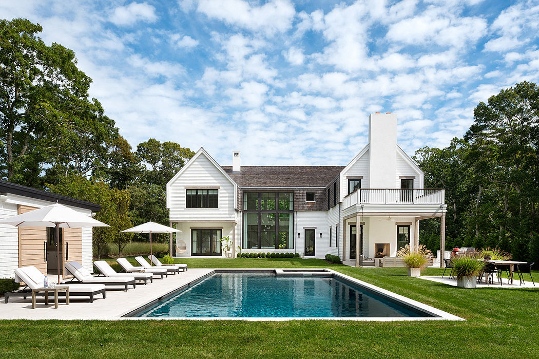 Expansive white modern farmhouse with large windows, balcony, and pool in lush green landscape.
