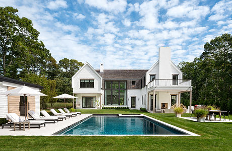 Expansive white modern farmhouse with large windows, balcony, and pool in lush green landscape.