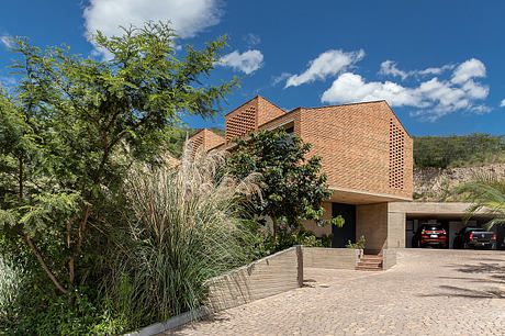 A modern brick house with a tiered roof, surrounded by lush vegetation and a paved driveway.