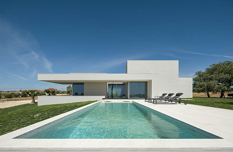 Modern white house with a large swimming pool and lounge chairs on a grassy lawn against a blue sky.