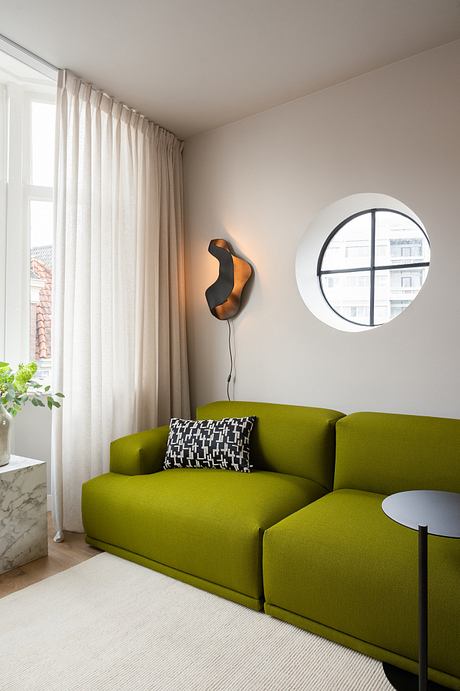 Modern living room with green sofa, round window, and abstract wall sconce.