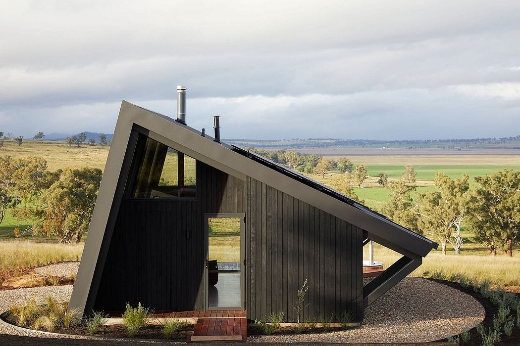 Small triangular cabin with black wood siding, sloped roof, and windows against rural landscape.
