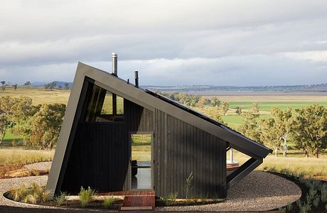 Small triangular cabin with black wood siding, sloped roof, and windows against rural landscape.