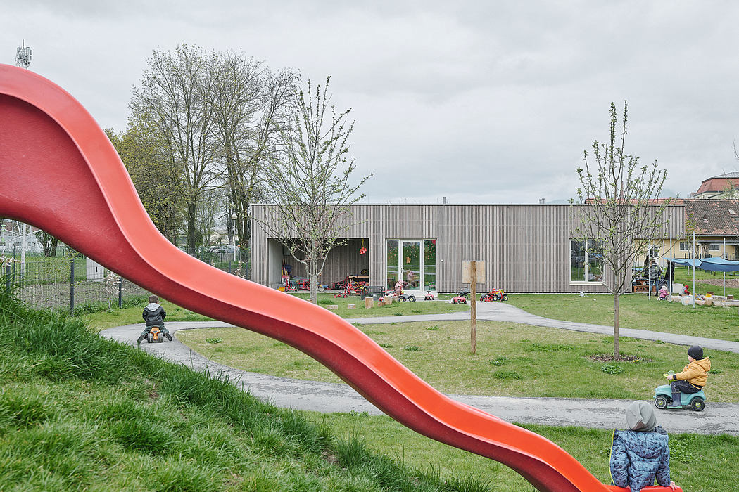 A modern playground with a large red slide, surrounded by trees and a contemporary building.