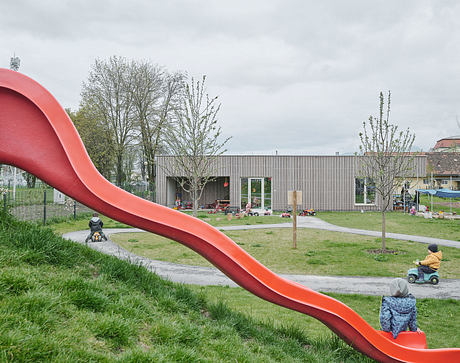 A modern playground with a large red slide, surrounded by trees and a contemporary building.