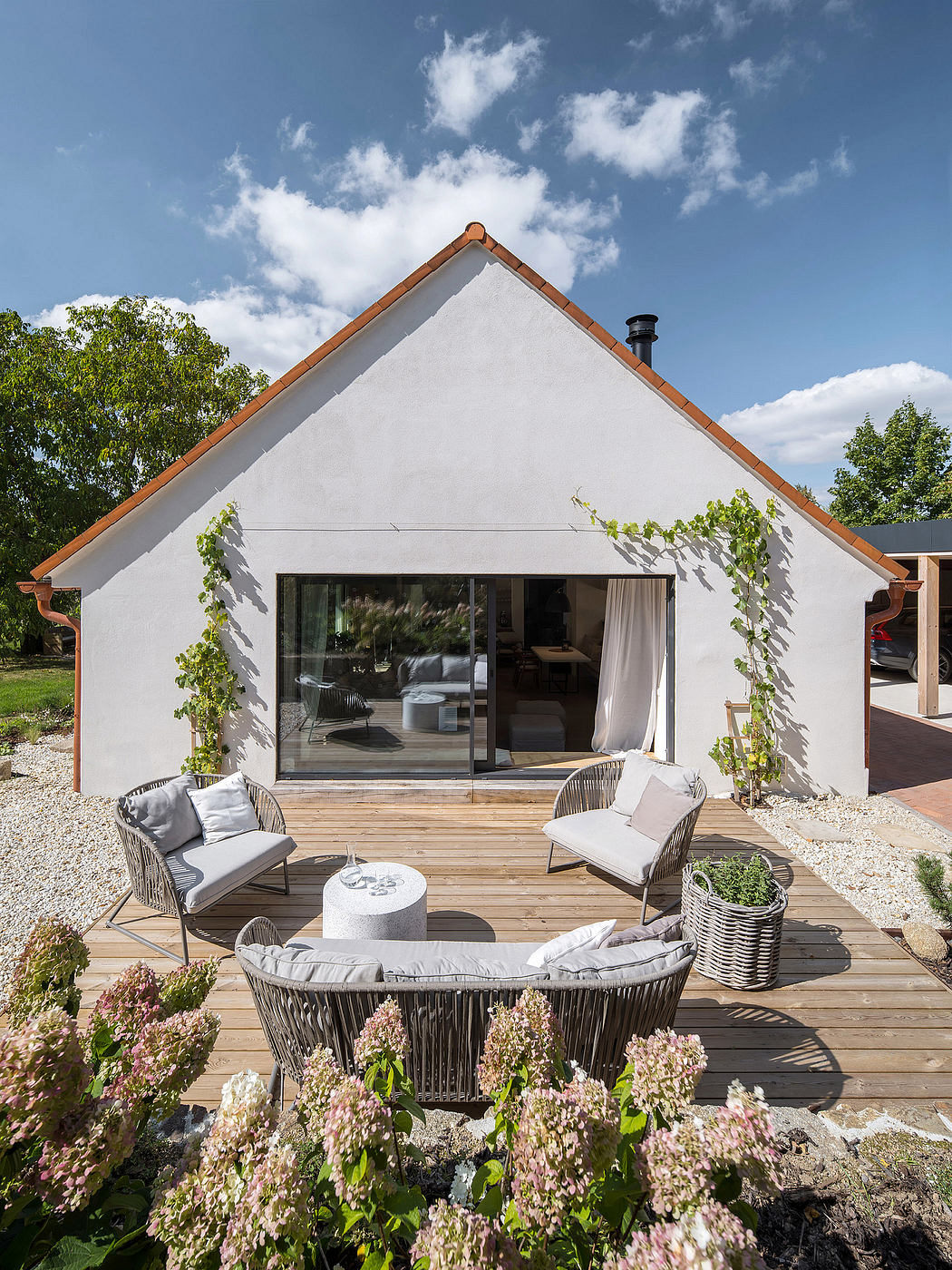 A modern white home with a large wooden deck, wicker furniture, and a floral garden.