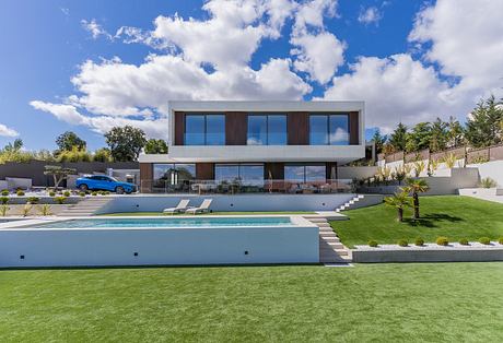 Modern, two-story house with sleek glass facade, palm trees, and a large pool surrounded by lush greenery.