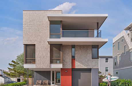 A modern, two-story house with shingle siding, glass balconies, and a vibrant red door.