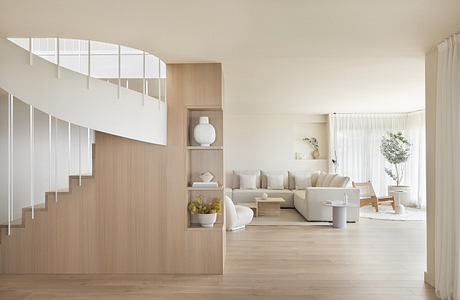 Modern, minimalist living room with curved staircase, neutral tones, and natural light from large windows.