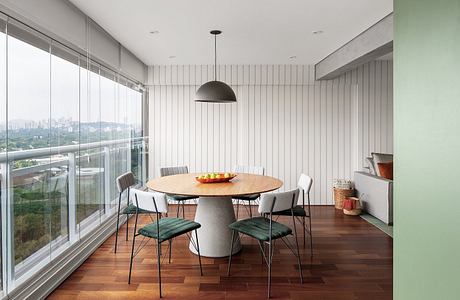 Modern dining room with round concrete table, green upholstered chairs, and large windows overlooking a city skyline.