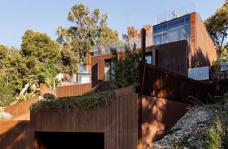 Modern multilevel building with wooden facade and glass panels, surrounded by lush greenery.