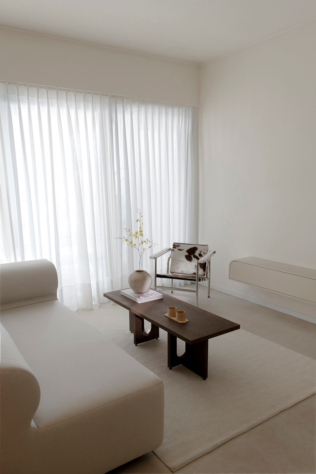 Minimalist living room with white curtains, wooden coffee table, and neutral furnishings.