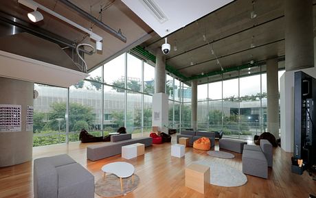 Modern open-plan office space with glass walls, wooden floors, and comfortable seating arrangements.