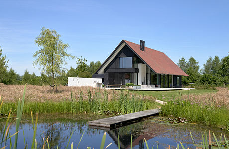 A modern chalet-style house with a red tiled roof, large windows, and a tranquil pond in the foreground.