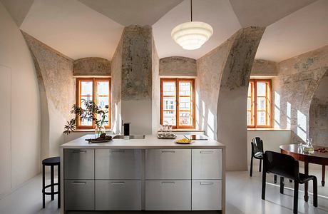 A modern, minimalist kitchen with stone arched walls, large windows, and sleek gray cabinets.