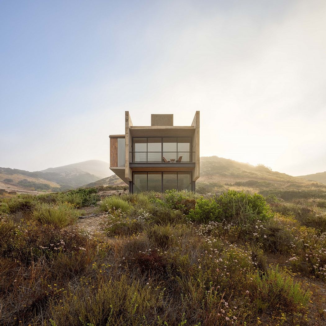 A modernist, glass-walled house perched atop a hill amid lush, diverse vegetation.