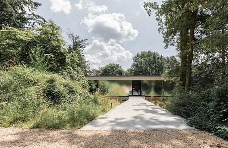 A simple, glass-fronted building with a flat roof set in a lush, wooded landscape.