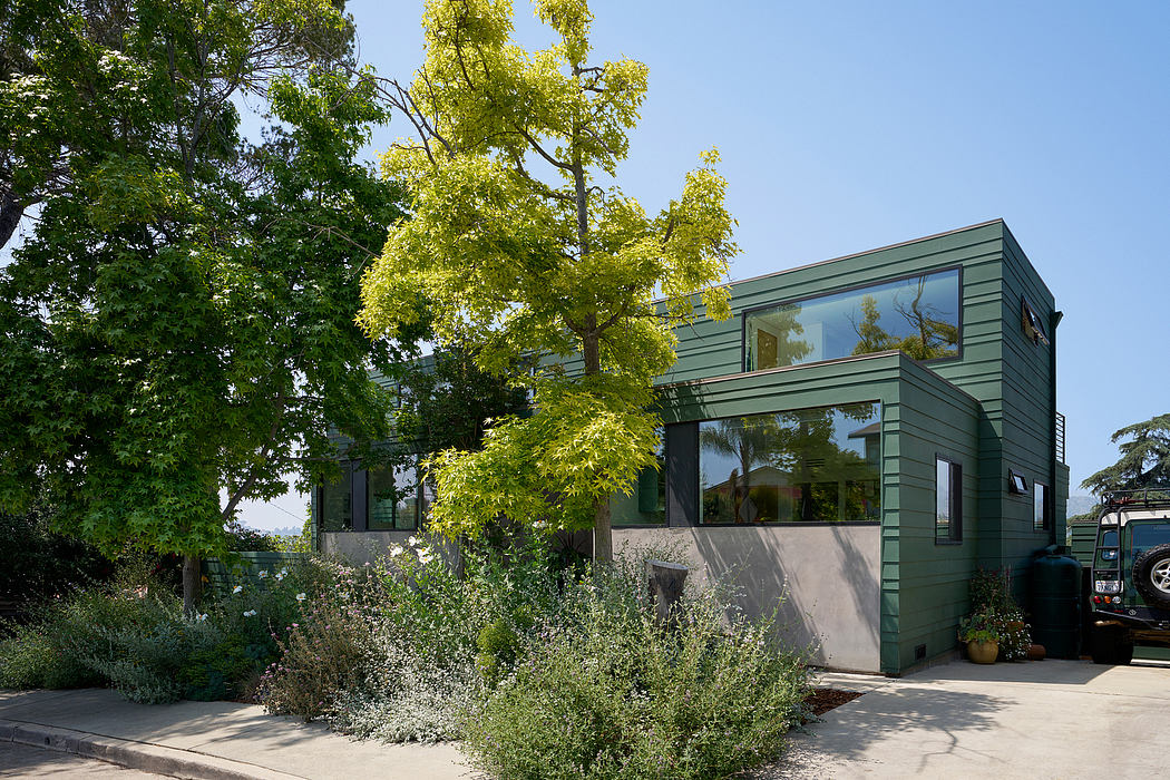 Modern green-painted home with large windows, surrounded by lush landscaping and trees.