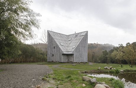 A modern angular wooden structure surrounded by a pond and lush vegetation.