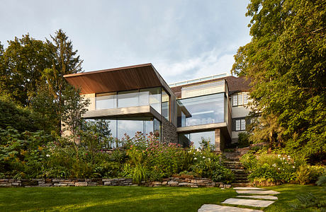 A modern, glass-enclosed home with a wooden roof set in a lush, landscaped garden.