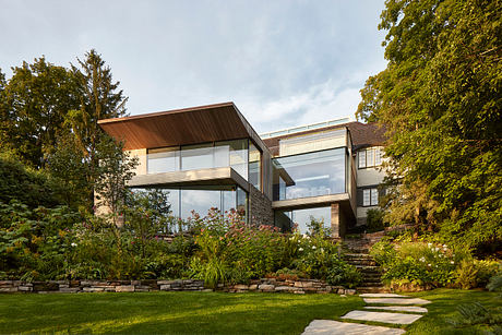 A modern, glass-enclosed home with a wooden roof set in a lush, landscaped garden.