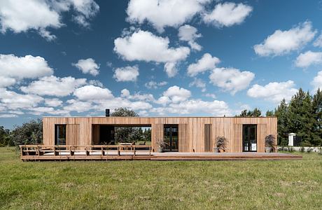 A modern, wooden cabin-style structure with a large deck overlooking a grassy field and pine trees.