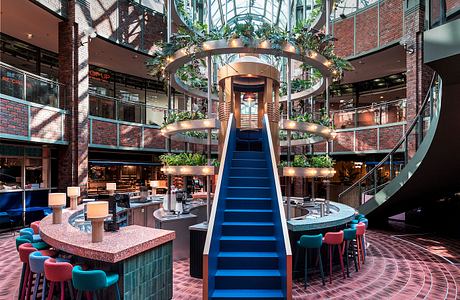 Vibrant modern atrium with geometric staircase, hanging plants, and colorful furnishings.