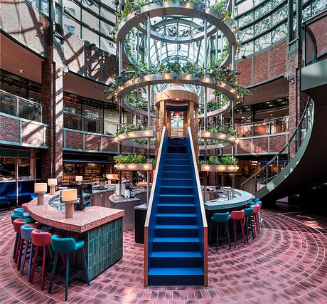 Vibrant modern atrium with geometric staircase, hanging plants, and colorful furnishings.