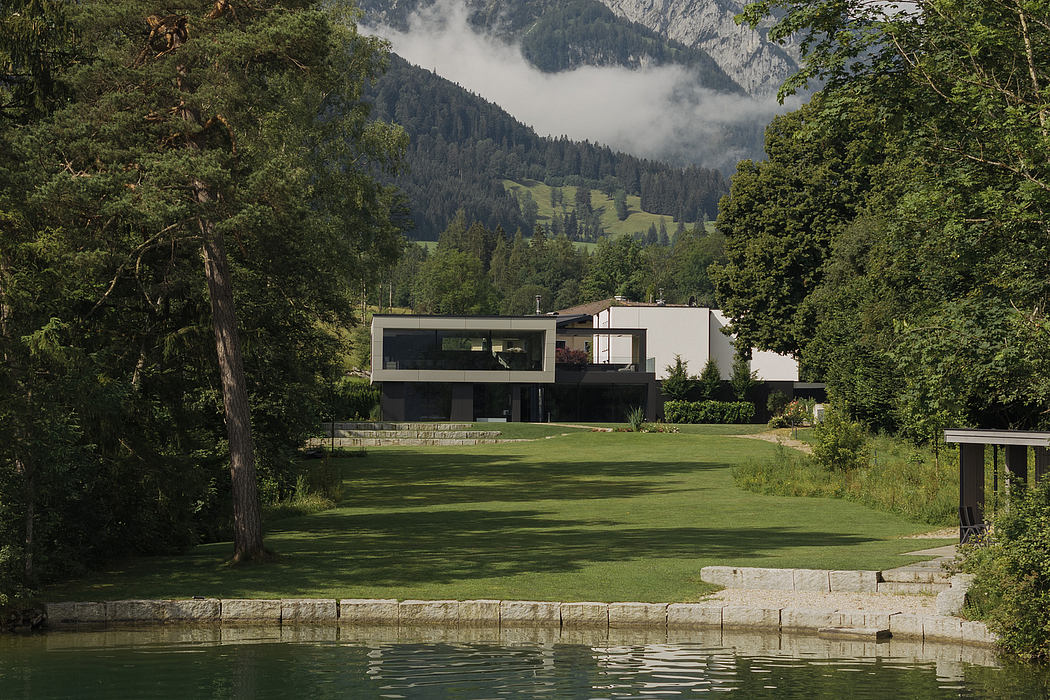 A modern, glass-walled house overlooking a lush, grassy meadow and distant mountains.