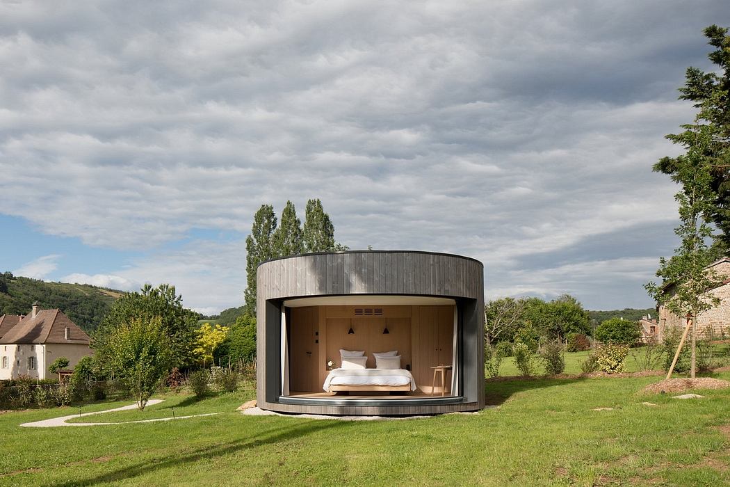 A circular wooden cabin with a curved roof, surrounded by a grassy field and lush greenery.