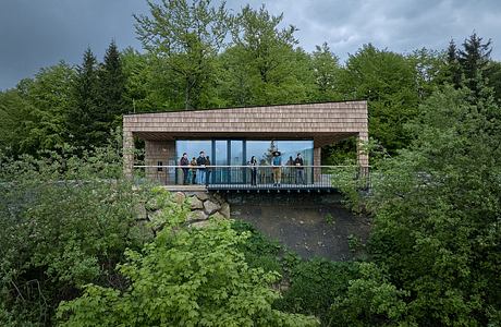 A modern wooden structure with large windows overlooks a lush, forested landscape.