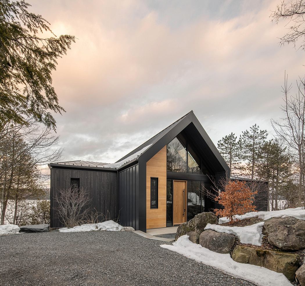 A modern, angular cabin with black siding, wood accents, and expansive windows overlooking a snowy landscape.