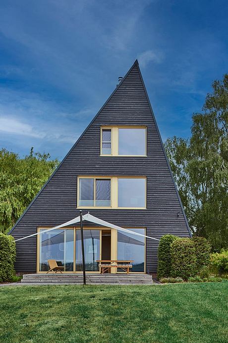 A modern, A-frame house with large windows, a glass entryway, and a surrounding garden.