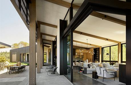 Contemporary open-plan living area with exposed wood beams, large windows, and outdoor dining.