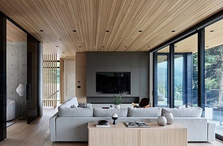 Large, modern living room with wood-paneled ceiling, gray sofa, and glass walls overlooking nature.