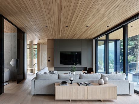 Large, modern living room with wood-paneled ceiling, gray sofa, and glass walls overlooking nature.
