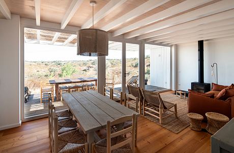 A cozy, rustic living and dining space with wooden beams, large windows, and natural furnishings.