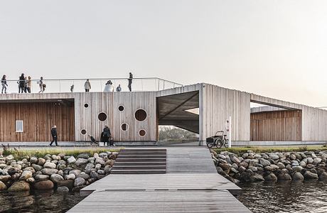 Wooden structure with arched openings and circular cutouts, surrounded by rocks and water.