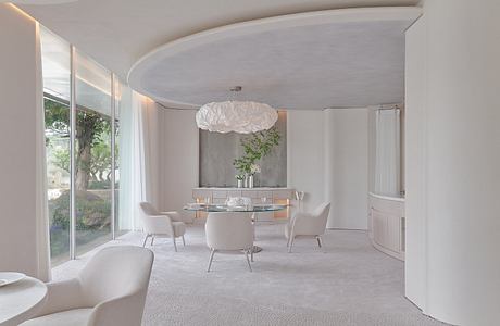 A spacious, minimalist dining area with curved walls, a cloud-like light fixture, and glass doors overlooking greenery.