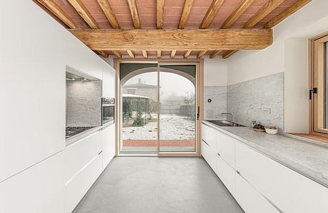 A modern kitchen with a rustic wood ceiling, white cabinets, and a large arched window overlooking an outdoor yard.