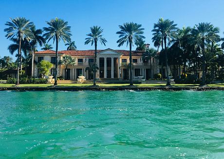 A grand, Spanish-style mansion with a towering facade and ornate balconies, surrounded by lush palm trees and turquoise waters.