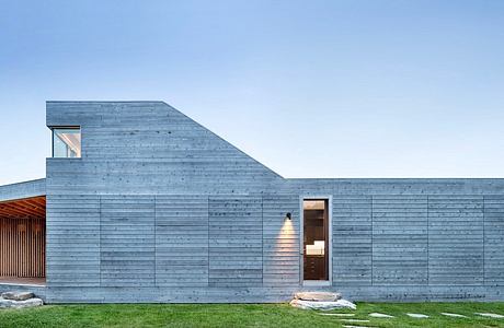 A modern, angular building with a slate-gray exterior and large windows, set against a grassy landscape.