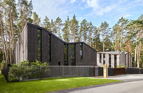 A modern, angular building with dark wood siding and large glass windows, surrounded by pine trees.