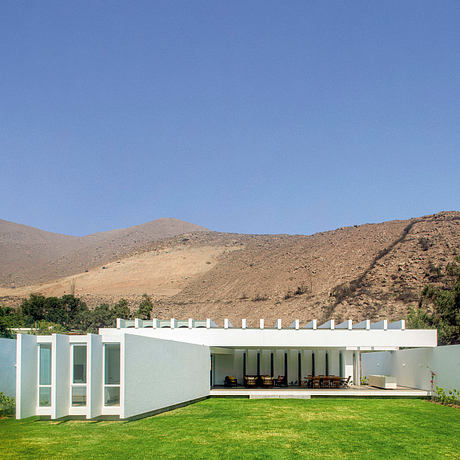 A modern, minimalist desert residence with an expansive patio and lush lawn surrounded by stark, arid hills.