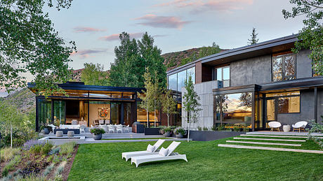 A modern, glass-enclosed home with a lush green lawn, patio, and sweeping mountain views.