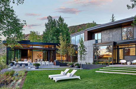 A modern, glass-enclosed home with a lush green lawn, patio, and sweeping mountain views.