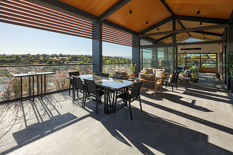 A modern patio with wooden beams, glass walls, and a dining area overlooking a scenic view.